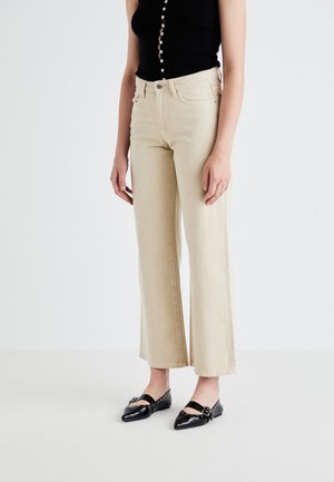 Jeans beige a vita alta e gamba larga con design classico a cinque tasche, abbinati a scarpe nere basse con cinturino e fibbia decorativa.