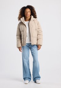 Giubbotto puffer beige con cappuccio, chiusura a zip e bottoni a pressione; indossato sopra una maglietta leggera e abbinato a jeans a gamba larga blu e sneaker bianche.