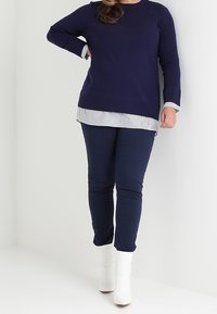 Navy-Pullover mit langen Ärmeln, übereinander getragen mit einem weiß-gestreiften Hemd in Navy, kombiniert mit navyfarbenen Hosen und weißen Stiefeletten.