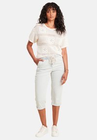 Gehaakte crèmekleurige trui met een open haakpatroon, gecombineerd met lichtblauwe joggingbroeken. Model draagt witte sneakers en poseert met één hand op de heup.