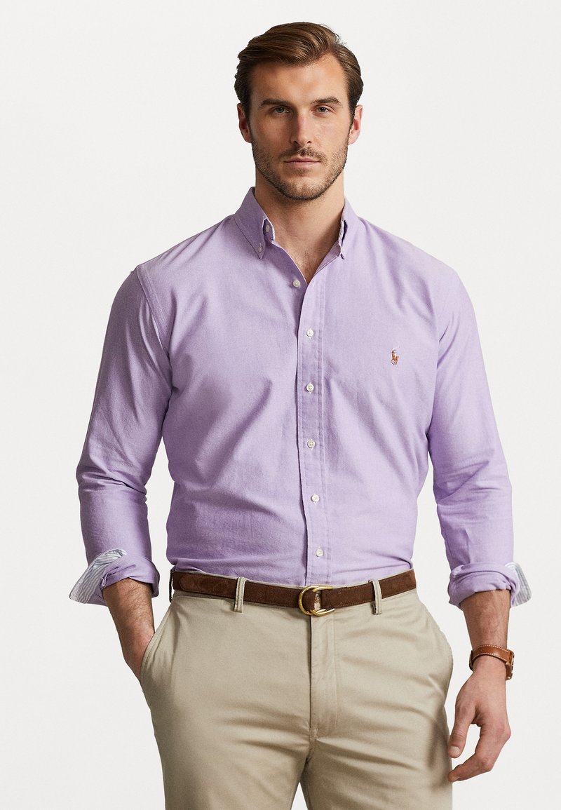 Λιλά πουκάμισο με κρίκου που κυλάει, φτιαγμένο από βαμβάκι. Καφέ δερμάτινη ζώνη με χρυσό κούμπωμα; φορεμένο με μπεζ παντελόνι.