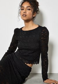 Svart spets långärmad topp med en vågig nederkant och puffärmar. Det blommiga mönstret skapar en texturerad look, i kombination med en figursydd silhuett.