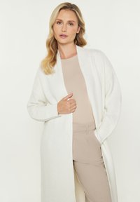 Cardigan lung, deschis în față, din material tricotat alb, cu un model texturat. Modelul poartă un top și pantaloni bej, cu mâneci largi și fără închideri.