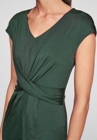 Grön omslagsklänning med v-ringning, korta ärmar och skrynkligt tyg vid midjan, tillverkad av mjukt, elastiskt material.