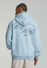 Lys blå hettegenser med et stort brodert design med sølvfiner og stjerner. Avslappet passform med snor i hetta og elastisk liv.