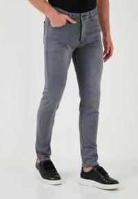 Jeans de denim gris con un corte slim, que presentan un diseño clásico de cinco bolsillos y un ligero desgaste, combinados con zapatillas negras tipo slip-on.