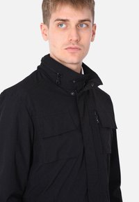 Giacca nera realizzata in tessuto resistente, con colletto alto, chiusura con zip e due grandi tasche sul petto con patte. Design minimalista.