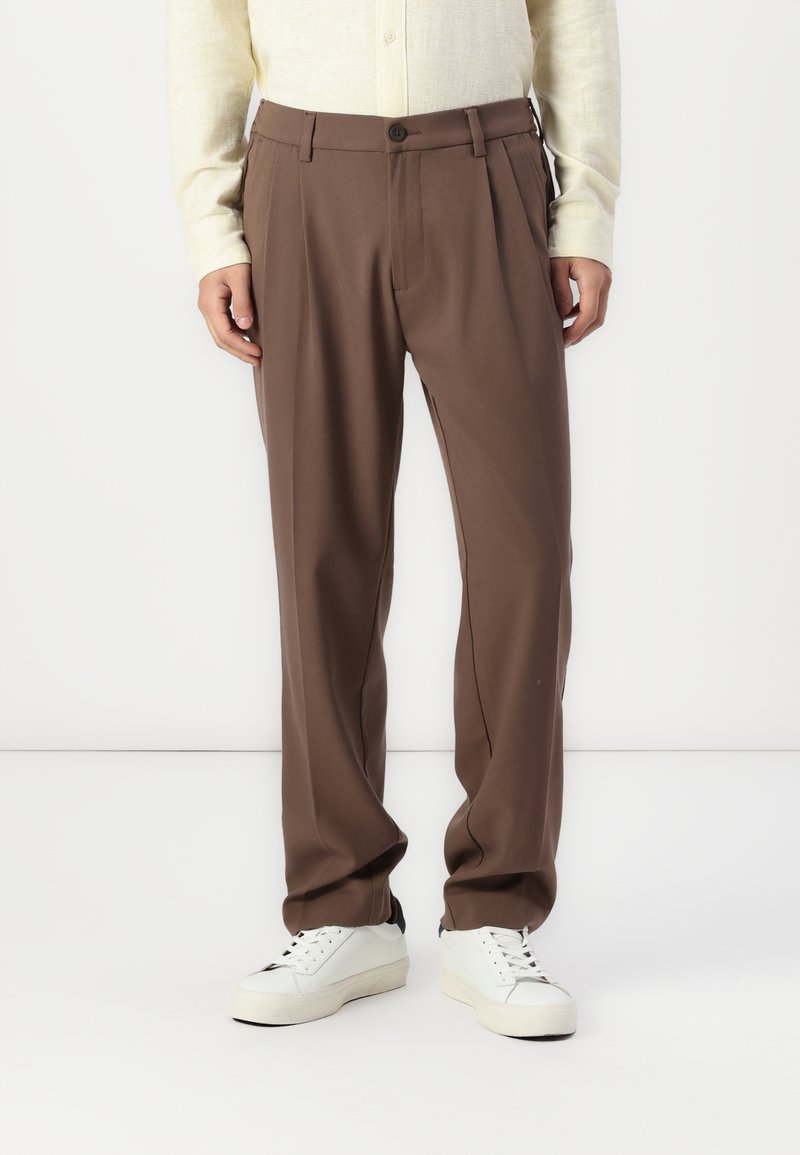 Brown pleated trousers with a straight fit, made from smooth fabric, featuring a button closure and side pockets. Styled with white sneakers.
