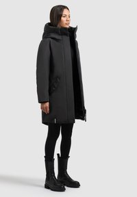 Mustmoodne veekindel parka kõrge kaelusega, küljetaskudega ja pika, sirge lõikega. Kantud mustade retuuside ja põlvi katvate mustade põlvikute kohal.