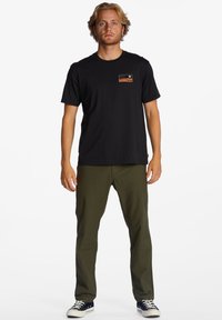 Sort bomulds T-shirt med korte ærmer og et lille grafisk motiv på forsiden, parret med olivengrønne bukser og marineblå sneakers.