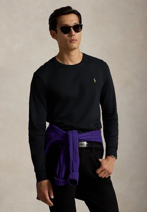 Mężczyzna ubrany w czarną koszulę z długim rękawem, czarne spodnie, okulary przeciwsłoneczne, z fioletowym swetrem związanym wokół talii na neutralnym tle.