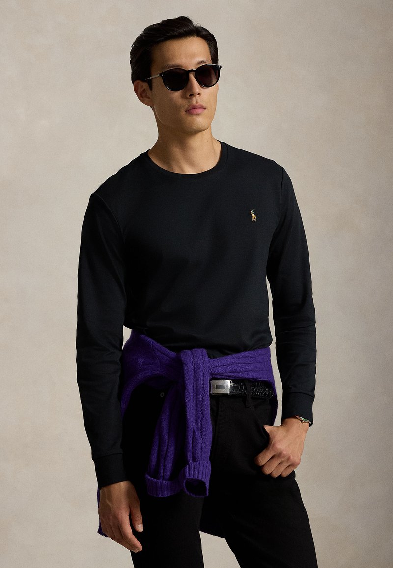 Mand iført sort langærmet skjorte, sorte bukser, solbriller og en lilla sweater bundet om livet mod en neutral baggrund.