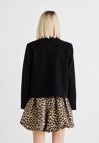 Melnā apgrieztā jaka ar garām piedurknēm, kas saskaņota ar plecu svārkiem ar leopardu rakstu, kuros ir zelta un melna krāsu shēma.