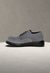 Chaussure habillée en suède gris à lacets avec une semelle noire, présentée sur une surface claire contre un fond beige uni.