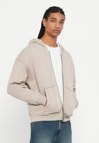 Sweatshirt bege com fecho, capuz ajustável, bolsos frontais e punhos canelados, usado sobre uma camisa branca, combinado com jeans azuis.
