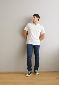 Weißes Kurzarmhemd mit Brusttasche, blaue Jeans und schwarz-weiße Sneakers. Steht an einer grauen Wand auf einem Holzboden.