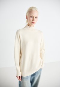 Cremfarbener Pullover mit hohem Kragen, gerippten Akzenten und asymmetrischem Saum. Hergestellt aus weichem Material und mit überschnittenen Schultern.