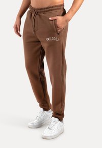Brune sweatpants lavet af blødt stof, med elastisk talje, sidelommer og hvid "SMILODOX" tekst. Båret med hvide sneakers.