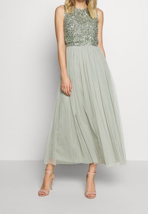 Vrouw draagt een mouwloze, lichtgroene, met pailletten versierde topjurk met een vloeiende, geplooide rok en nudekleurige hoge enkelband sandalen.