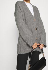 Grå virkade kofta med V-hals, stora knappar och överdimensionerade ärmar. Texturerat stickat tyg och ribbade detaljer vid ärmslut och nederkant.