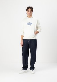 Sudadera crema con logo azul de "Guess Jeans", con bolsillo frontal. Combinada con jeans azul oscuro y zapatillas blancas, sobre un fondo blanco liso.