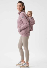 Felpa rosa con cappuccio e tasche frontali, dotata di un compartimento posteriore per trasportare un bambino. Abbinata a leggings beige e sneakers multicolori.