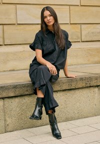 Femme aux longs cheveux portant un chemisier noir, un pantalon noir à volants et des bottes noires, assise sur un rebord en pierre contre un mur en pierre beige.