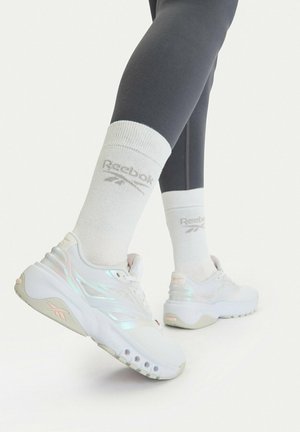 Nogi w czarnych legginsach, w białych skarpetkach Reebok i białych masywnych sneakersach z iryzującymi detalami na jasnym tle.