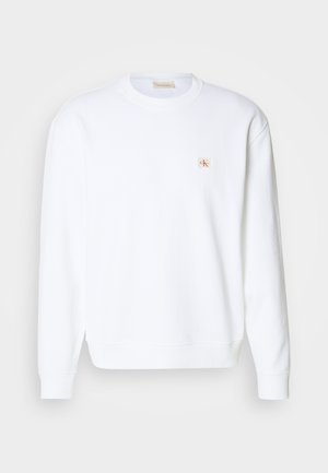 Fehér pulóver hosszú ujjakkal, kerek nyakkal és bordázott hüvelykujjakkal. Bal mellen egy kis piros és bézs logó található. Sima textúrájú.