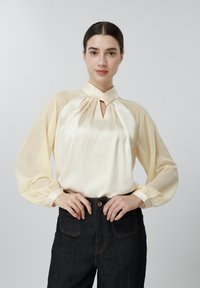 Ung kvinde iført en cremefarvet satinbluse med nøglehul-udskæring og puffede ærmer, kombineret med højtaljede mørke jeans, hænderne på hofterne.