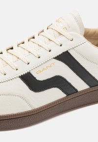Hvit lær sneakers med svarte bølgedesign detaljer. Har kremfargede snørebånd og beige innside. Gull "GANT" logo på siden. Gummisåle.