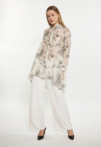 Blusa leggera con motivo floreale in beige e nero, dotata di bottoni sul davanti, maniche lunghe e un orlo posteriore più lungo. Abbinata a pantaloni bianchi.