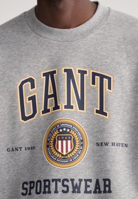 Šedá mikina s nápisem "GANT" v námořnické modři a žluté, s kulatým emblémem a textem "SPORTSWEAR". Kulatý výstřih a měkká textura.