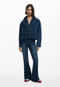 Denim takki, jossa on lyhennetty muotoilu ja suuret kaulukset, yhdistettynä leveälahkeisiin denim-housuihin. Molemmat vaatekappaleet ovat tummansinistä pesua.