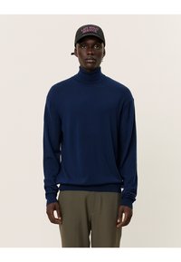 Marineblauer Rollkragenpullover aus weichem Stoff, mit langen Ärmeln und geripptem Kragen. Kombiniert mit grünen Hosen und einer schwarzen Mütze.