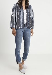 Veste oversized à motifs bleus avec un mélange de rayures et de motifs floraux, portée sur un haut blanc, associée à un jean déchiré et des chaussures blanches.