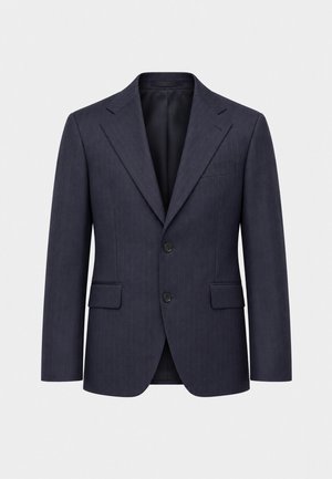 Un mannequin homme se tient devant un fond uni, vêtu d’un blazer bleu marine, d’un col roulé gris, d’un pantalon blanc et de mocassins beiges.