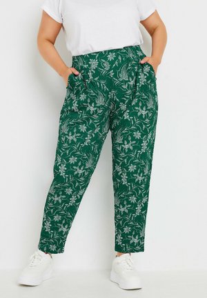 Pantalons verts à motifs avec un design floral, devant plissé, poches latérales et coupe fuselée, associés à des baskets blanches.