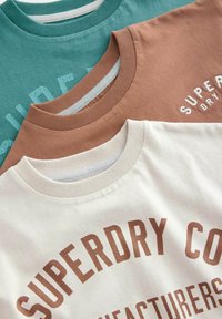 Tre T-shirts stablet: teal med prægede bogstaver, rustbrun med hvid tekst, og creme med brun trykt logo. Bomuldsstof, runde halsudskæringer.