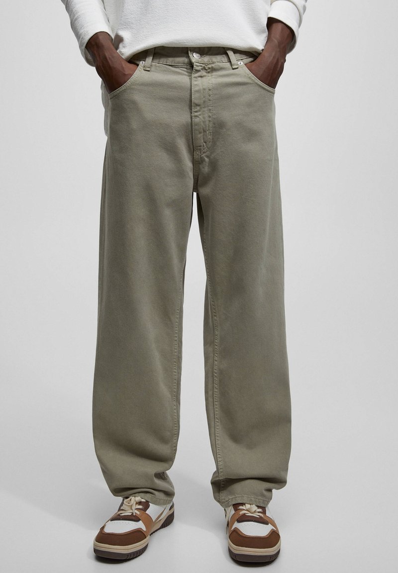 Personne portant un pantalon ample vert olive avec les mains dans les poches et des baskets blanches, marron et beige, debout sur un fond gris uni.