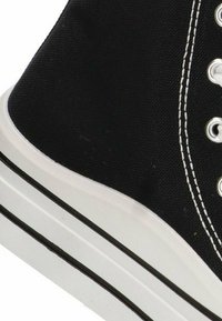 Zapatilla alta de lona negra con suela de goma blanca y franjas negras. Presenta ojales metálicos y costuras blancas en contraste.