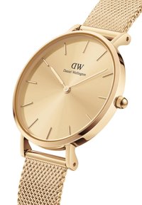 Montre dorée avec un bracelet en maille, présentant un cadran rond minimaliste et de fines repères horaires. Affiche "Daniel Wellington" sous le logo.