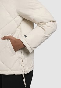 Chaqueta acolchada beige claro con un bolsillo lateral, detalle de cordones en el dobladillo y un puño de manga sólido con un pequeño parche negro.