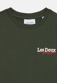 Tamsiai žalia medvilninė marškinėliai su apvalia iškirpte. Priekyje yra baltos ir raudonos spalvos užrašas "Les Deux Sporting Goods". Lygus paviršius.