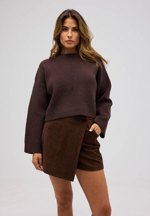 Kvinna med långt vågigt hår som bär en mörkbrun stickad tröja och asymmetriska bruna kordshorts, stående med en hand i fickan.