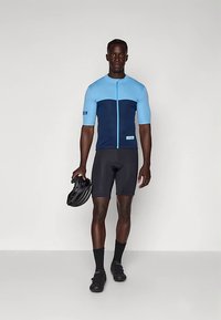 Kortärmad cykeltröja i ljusblått och marinblått, med blixtlås fram och märkning. Bärs med svarta shorts och skor, håller en svart hjälm.