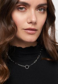 Metalhalstring med forlængede led i en sort sølv finish, med en cirkulær accent nær midten. Båret over en sort, ribbestrikket højhalset sweater.