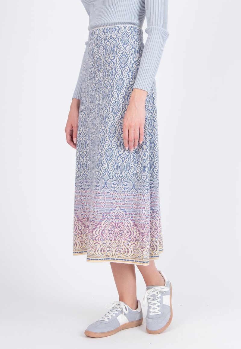 Kvinde iført en lang mønstret blå og pastel nederdel, en lyseblå ribbestrikket sweater og grå og hvide sneakers, stående på en hvid baggrund.