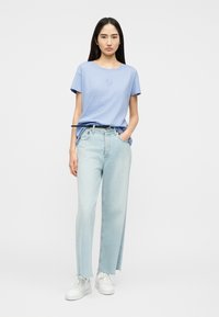 Νεαρή γυναίκα στέκεται με τα χέρια στις τσέπες, φοράει ένα ανοιχτό μπλε t-shirt, ξεβαμμένα τζιν με ίσια γραμμή και άσπρα αθλητικά παπούτσια σε λευκό φόντο.