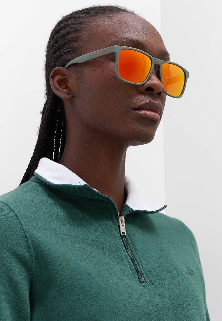 Zielone okulary przeciwsłoneczne z pomarańczowymi, lustrowymi soczewkami i matowym wykończeniem. Model ma na sobie zieloną bluzę zapinaną na zamek z białym kołnierzykiem.